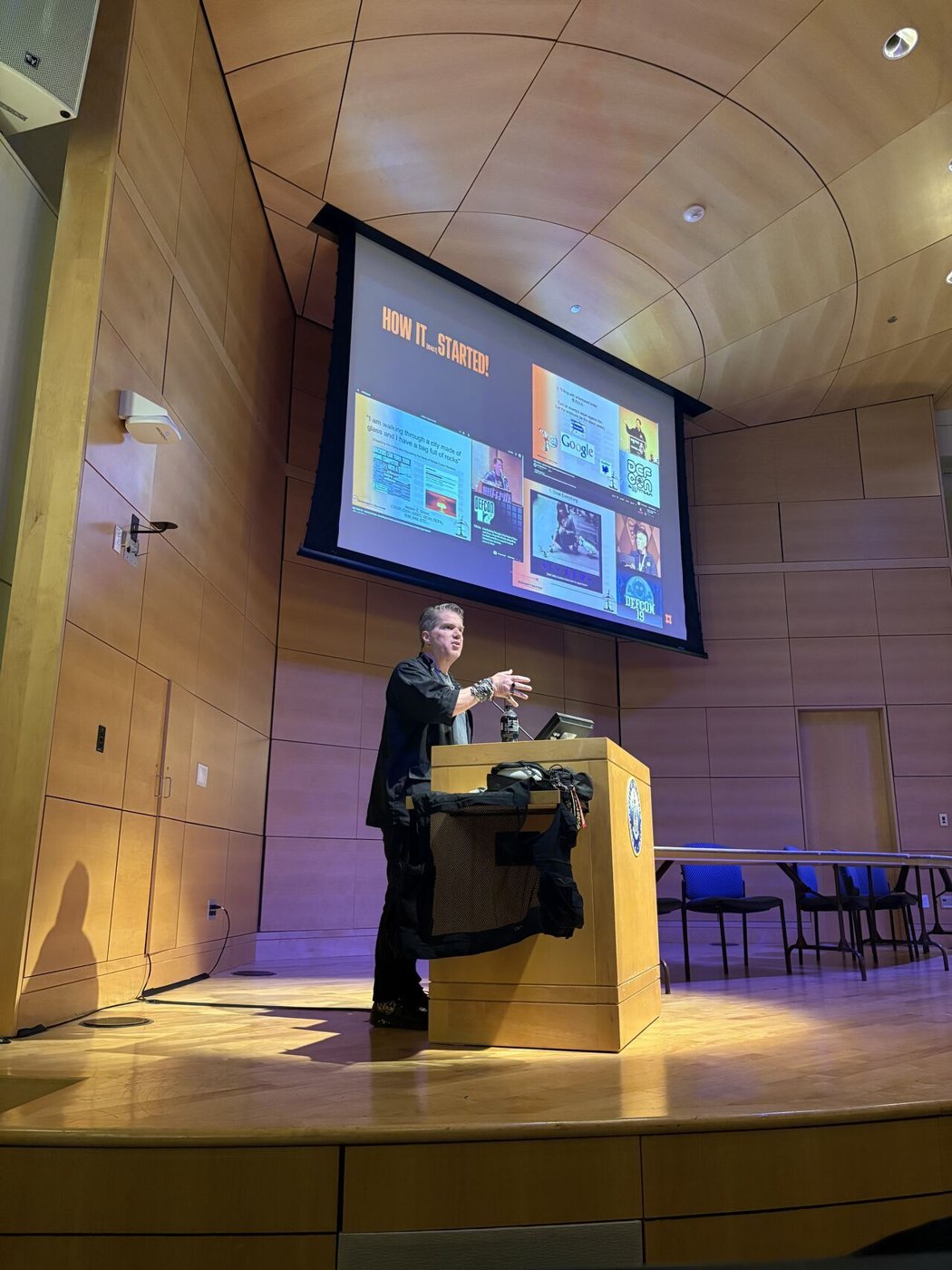 A speaker on stage at BSides Maine, gesturing in front of a large slide titled 'How it (really) started' collaged with early DEF CON and hacker-history images