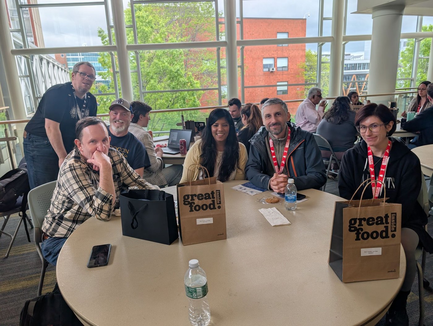 A group of attendees around a table at BSides Maine with boxed lunches in 'great food inside' bags, laptops open, more people and daylight windows behind them