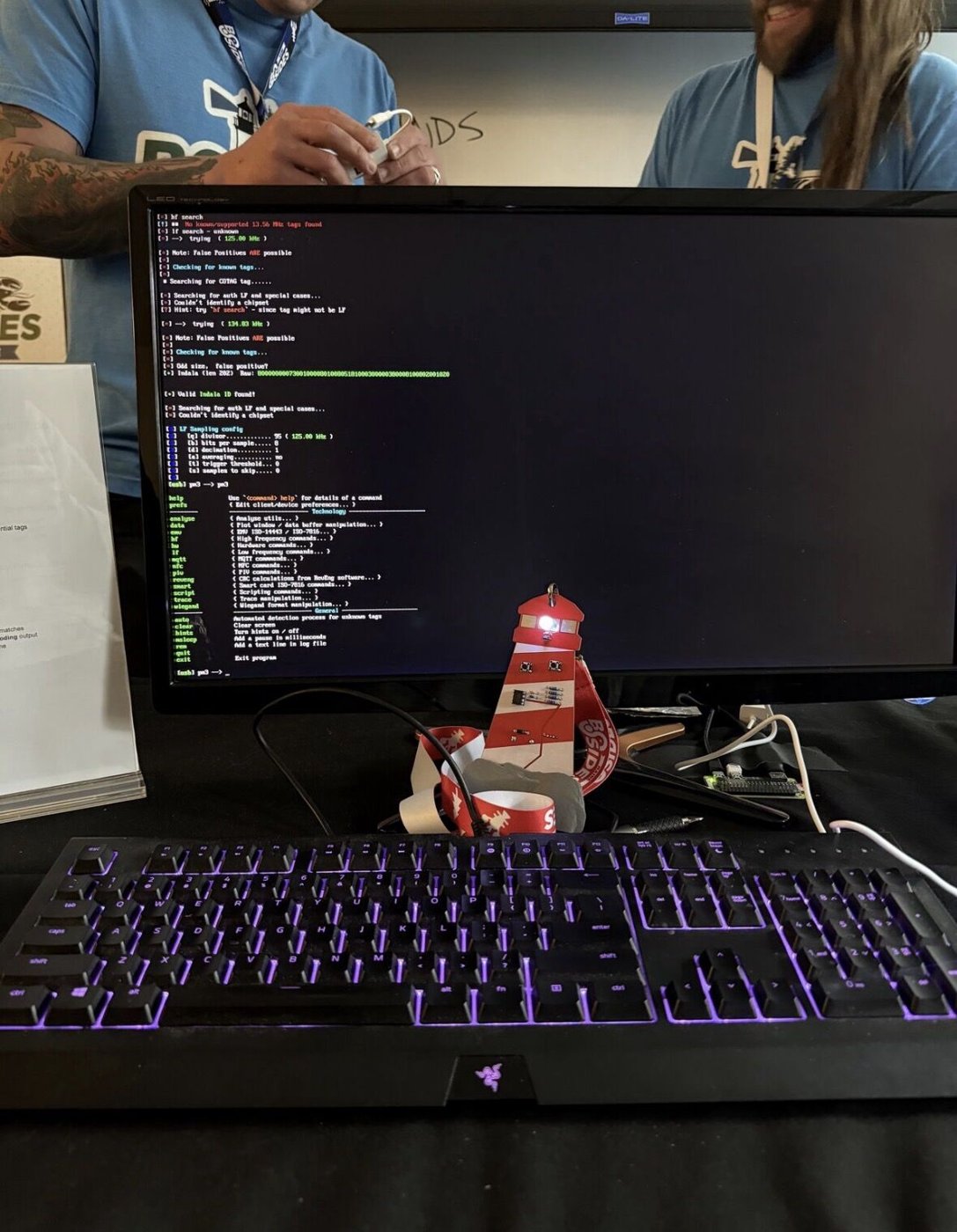A badge-hacking station: a monitor running a Proxmark RFID search in a terminal, a lit-up red lighthouse-shaped BSides badge in front of the keyboard, two people in BSides staff shirts working behind it