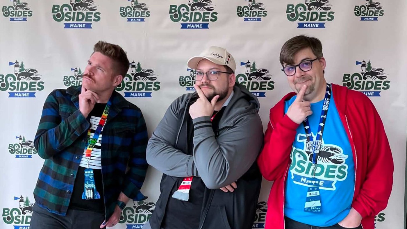 Three people posing in front of the BSides Maine step-and-repeat banner, each doing an exaggerated thoughtful chin-stroking pose