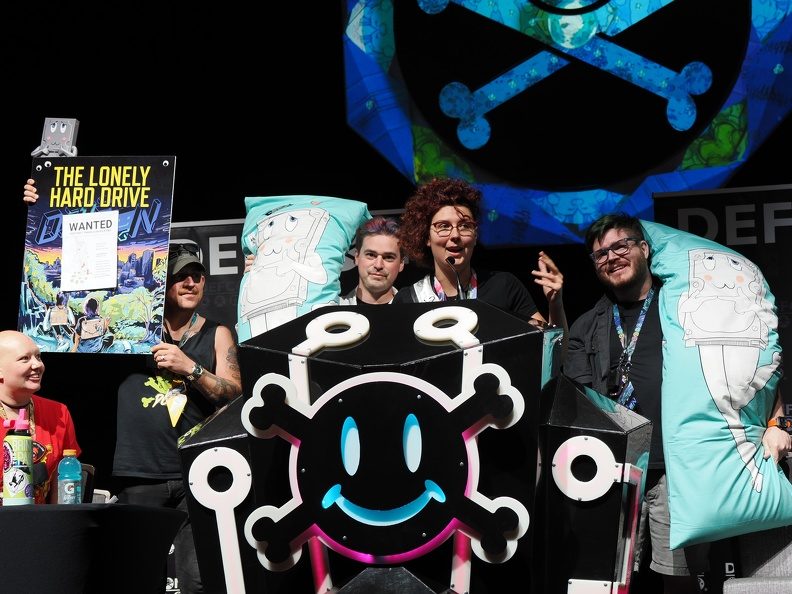 The Lonely Hard Drive crew on stage at DEF CON 31 with the contest's skull-and-crossbones hard-drive mascot and a "The Lonely Hard Drive" poster