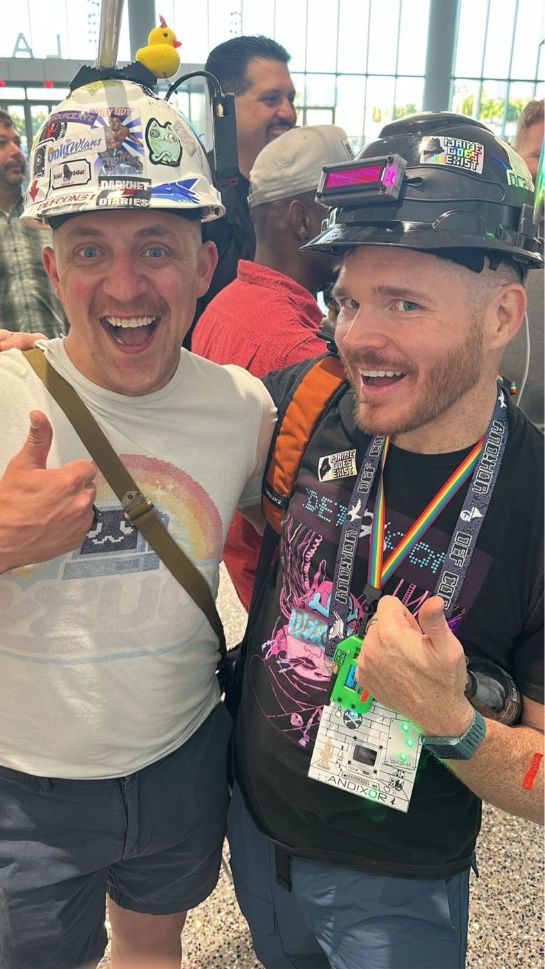 Two people grinning together in sticker-covered hard hats at DEF CON Linecon; one hat is topped with a rubber duck, the other shows a lit OLED display and a 'Maine Does Exist' sticker