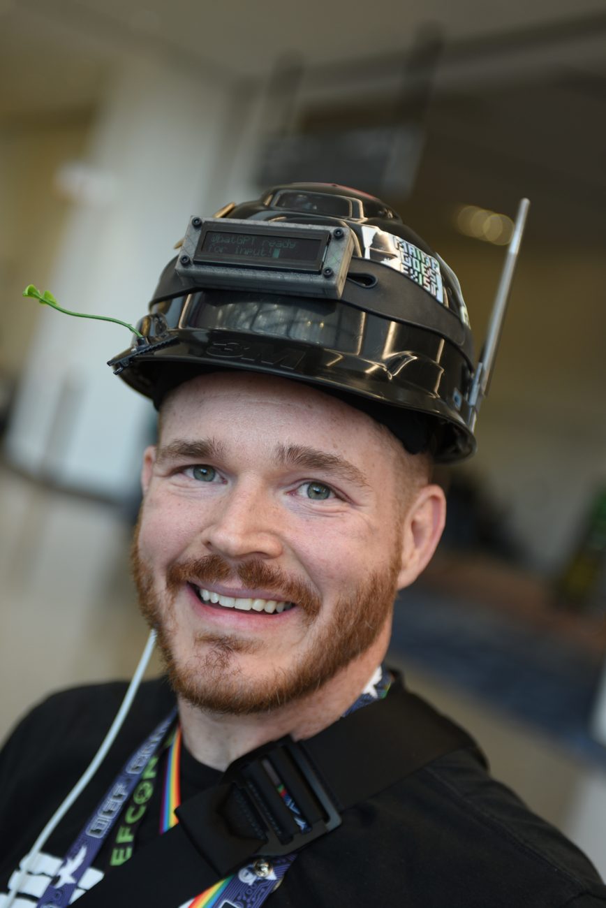 Me wearing HatGPT: a black hard hat with an OLED display reading 'HatGPT ready for input!', an antenna, and a 'Maine Does Exist' sticker