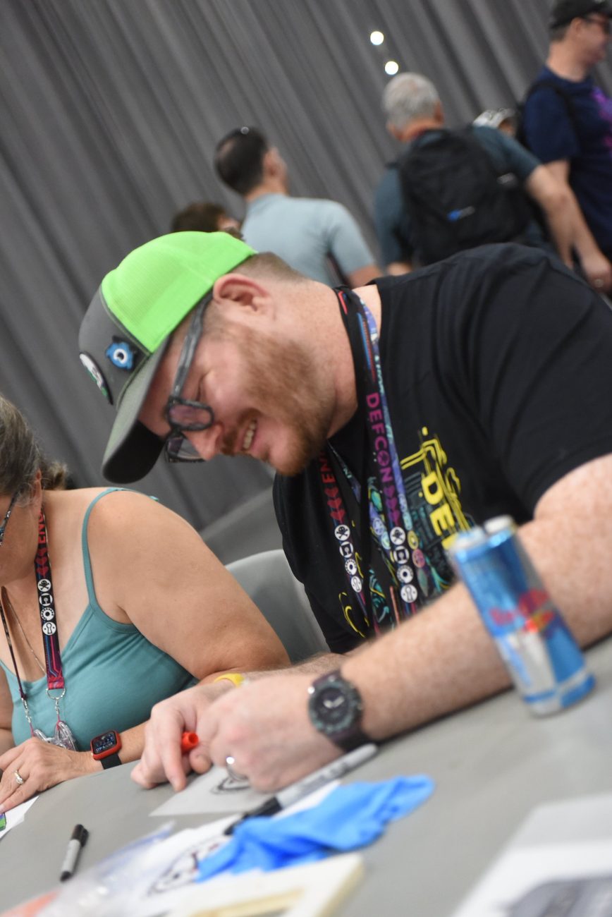 Me in a green cap soldering at a table in the DEF CON Maker Village, learning to build an SAO badge add-on