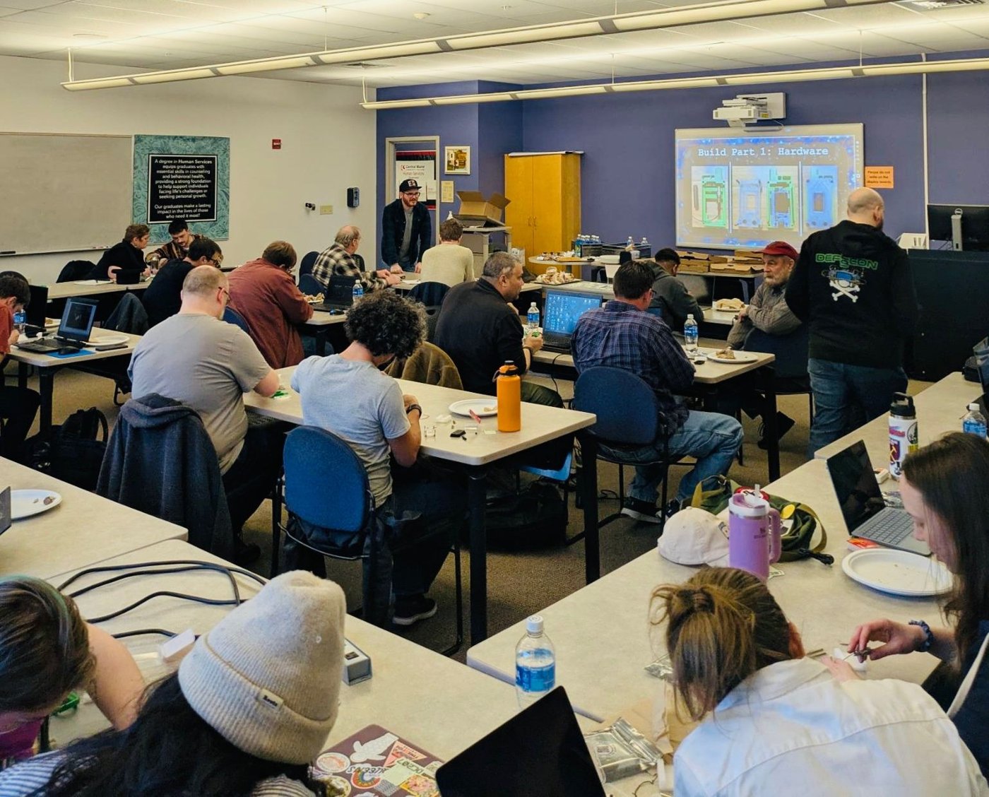 A DC207 hardware workshop in a classroom: attendees at long tables with laptops, components, and pizza, with a projected slide reading 'Build Part 1: Hardware'