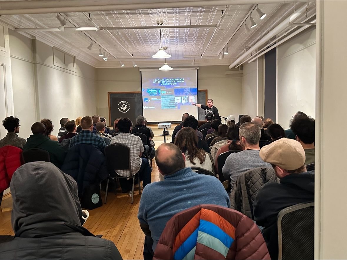 A packed DC207 meetup room in Portland with every seat full, a speaker gesturing at a projected slide reading 'I can't make cool hacking tools but I can get ChatGPT to turn my website into one'