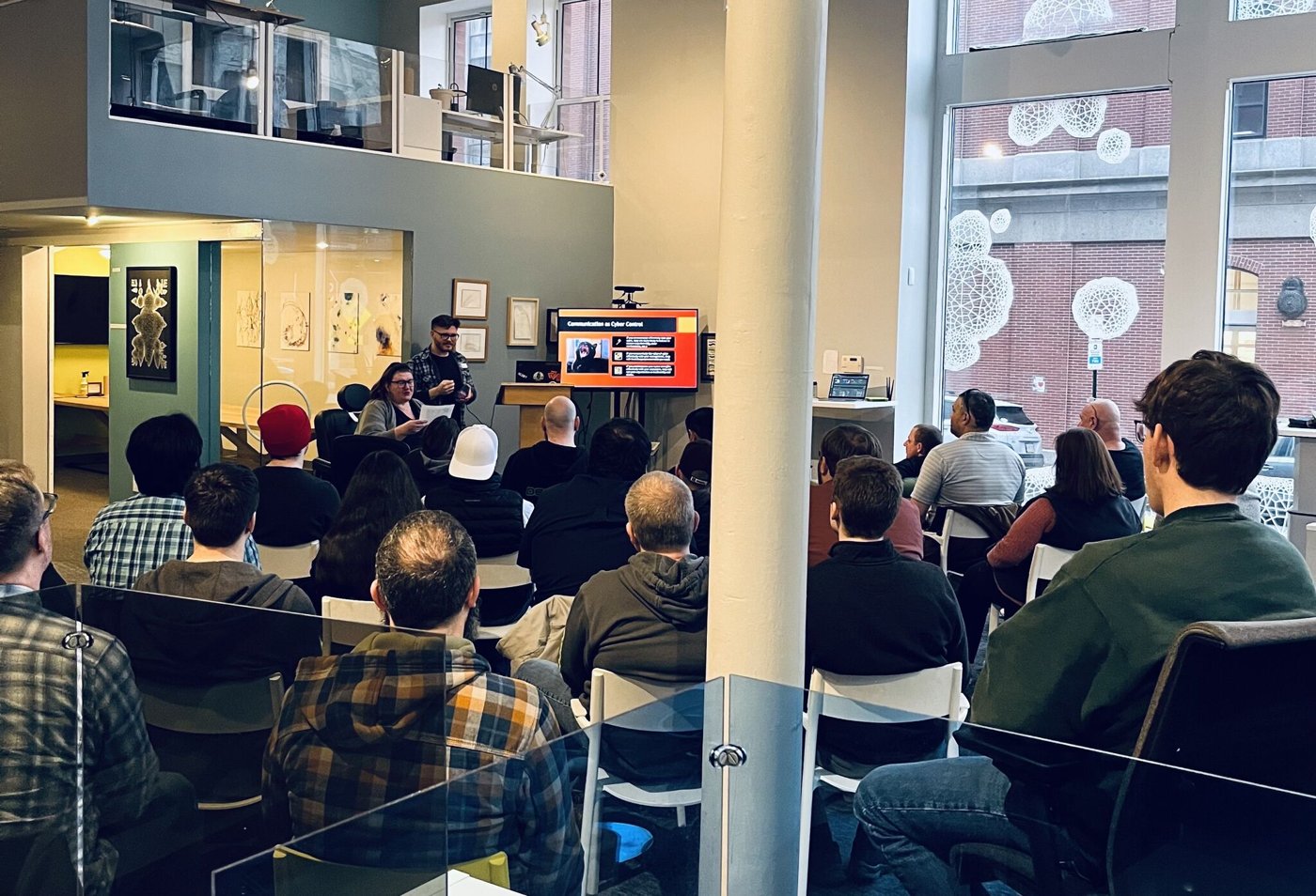 A DC207 lightning talk in a bright open venue: two presenters standing beside a screen reading 'Communication as Cyber Control' with a seated audience filling the room