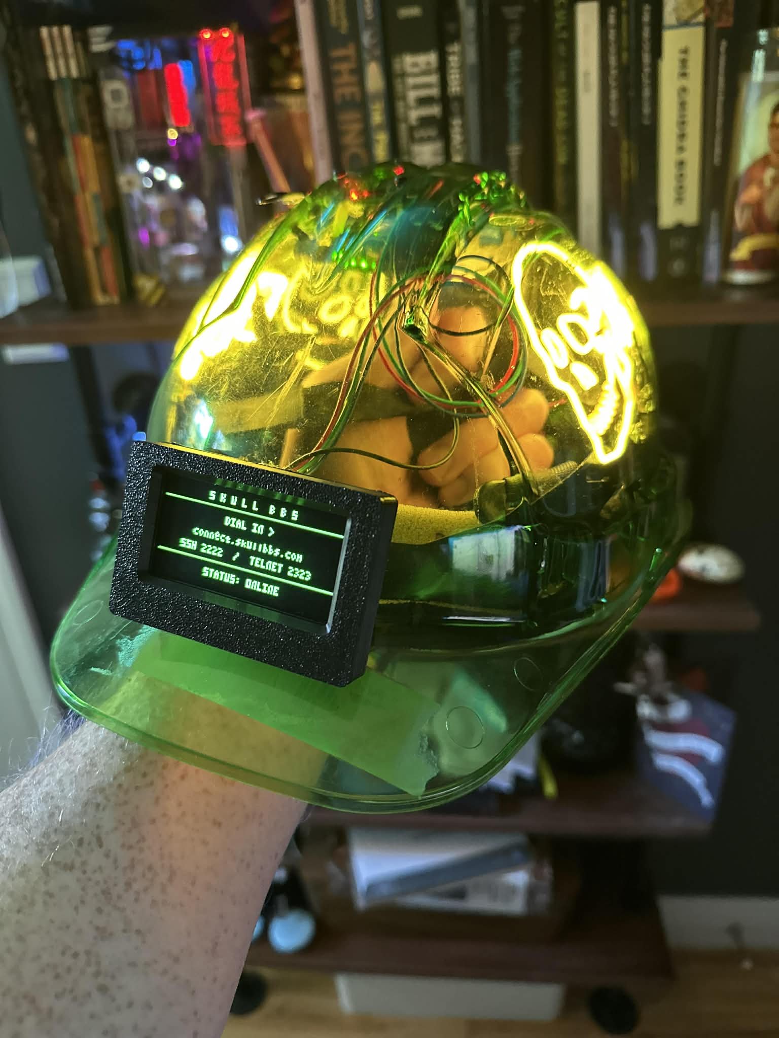 Close-up of the hard hat's brim-mounted OLED reading 'SKULL BBS / DIAL IN > / connect.skullbbs.com / SSH 2222 / TELNET 2323 / STATUS: ONLINE', with the glowing skull logo behind it.