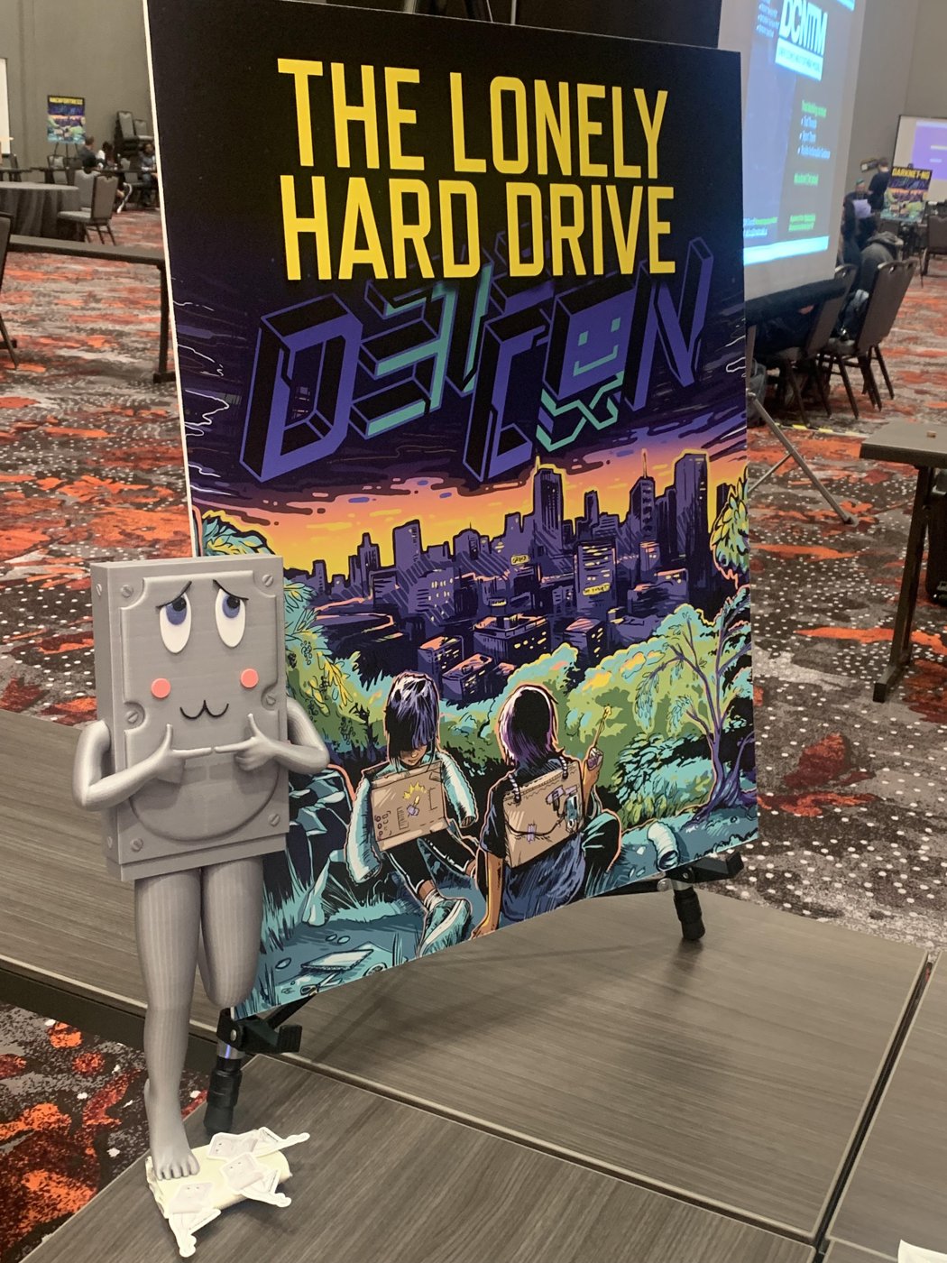 oh betty... Betty Pagefile, the shy 3D-printed grey hard-drive mascot with blushing cheeks, standing beside the Lonely Hard Drive DEF CON poster